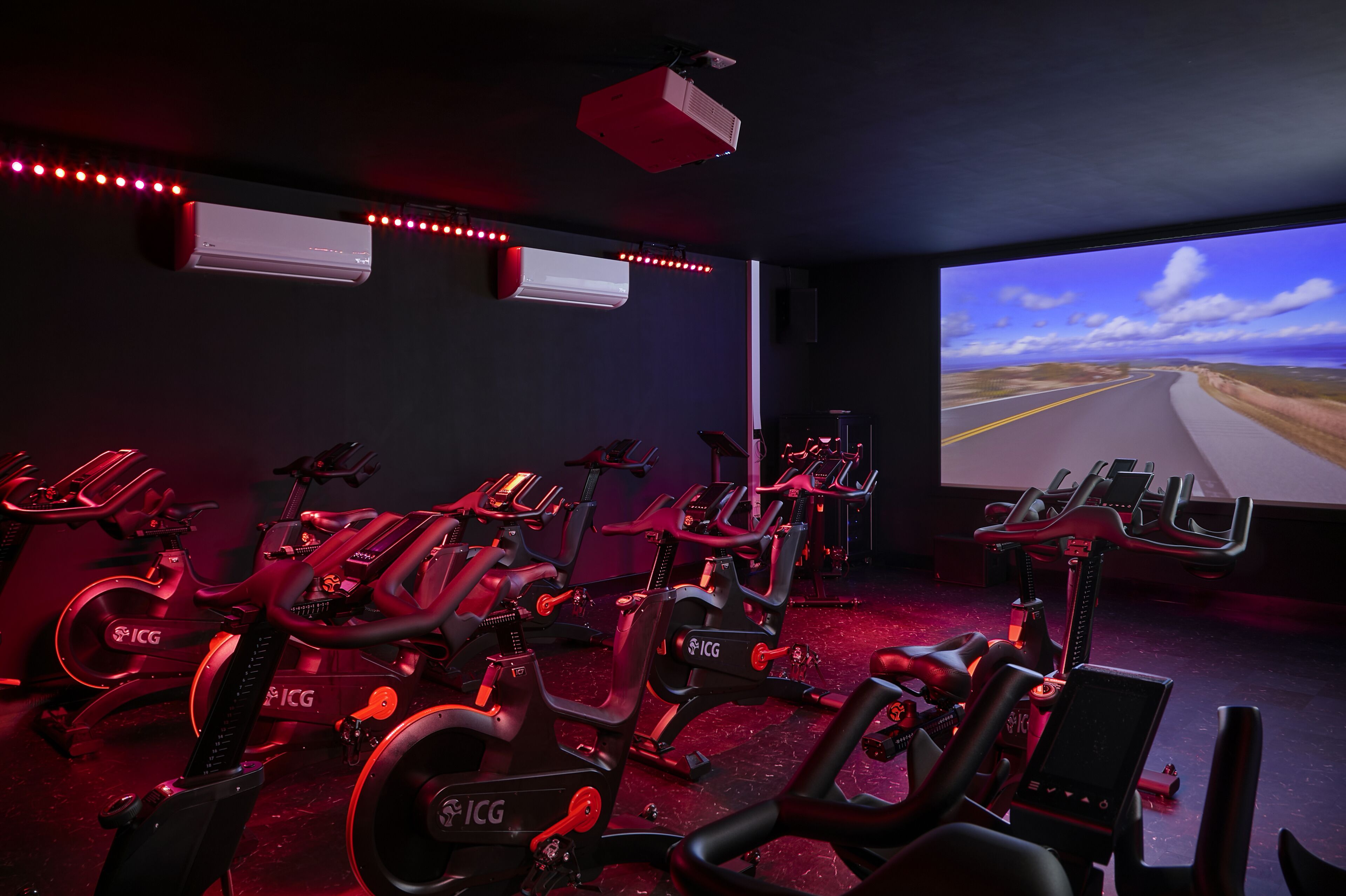 A gym with stationary bikes, air conditioning units, and a large screen displaying a scenic road view.