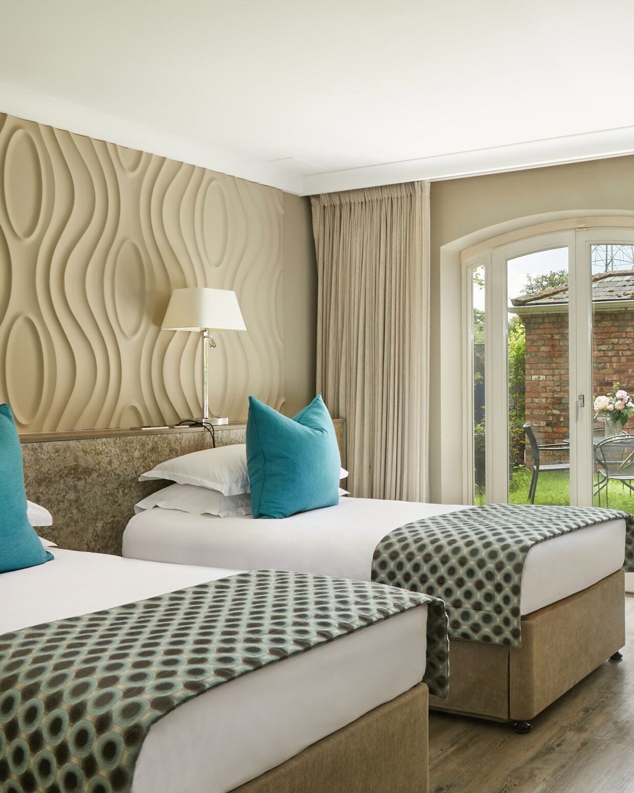 Ein Hotelzimmer mit zwei Betten, einem gemusterten Kopfteil, einer Lampe und Blick auf einen Garten durch eine Glastür.