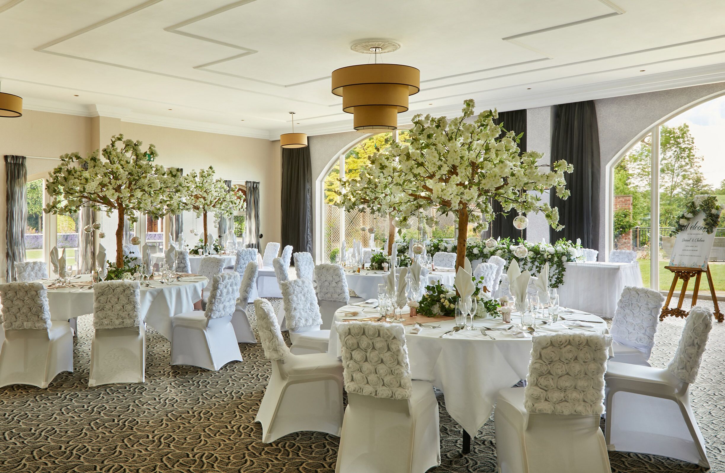 Una sala da banchetto con tavoli rotondi apparecchiati per un evento formale, con centri tavola floreali bianchi e un'elegante mise en place.