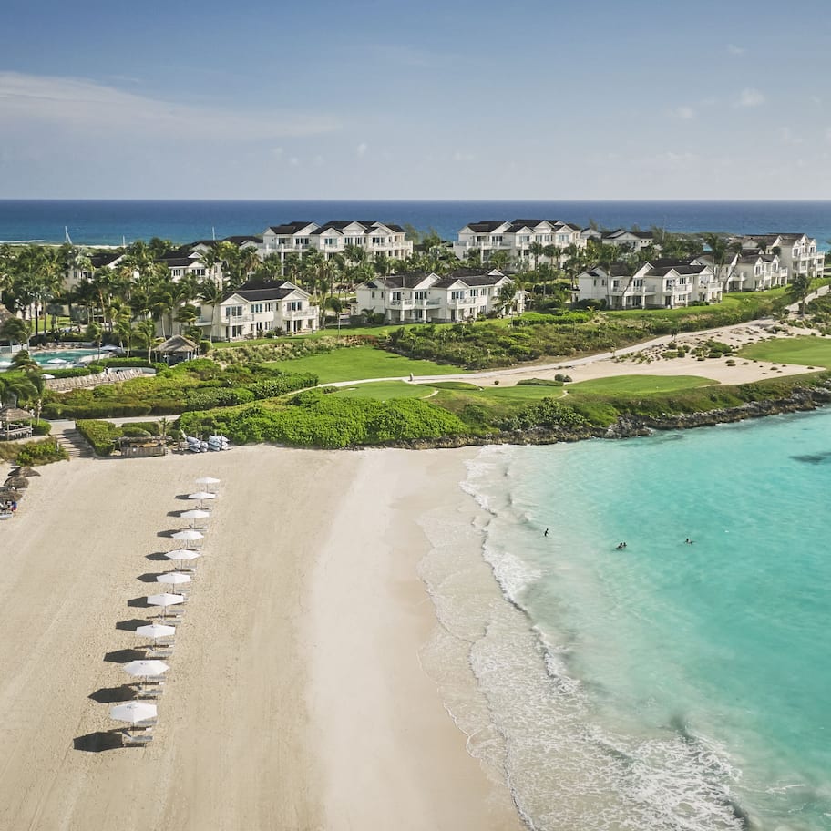 An aerial view of a coastal resort with beachfront villas, a sandy beach with umbrellas, and a clear view of the ocean.