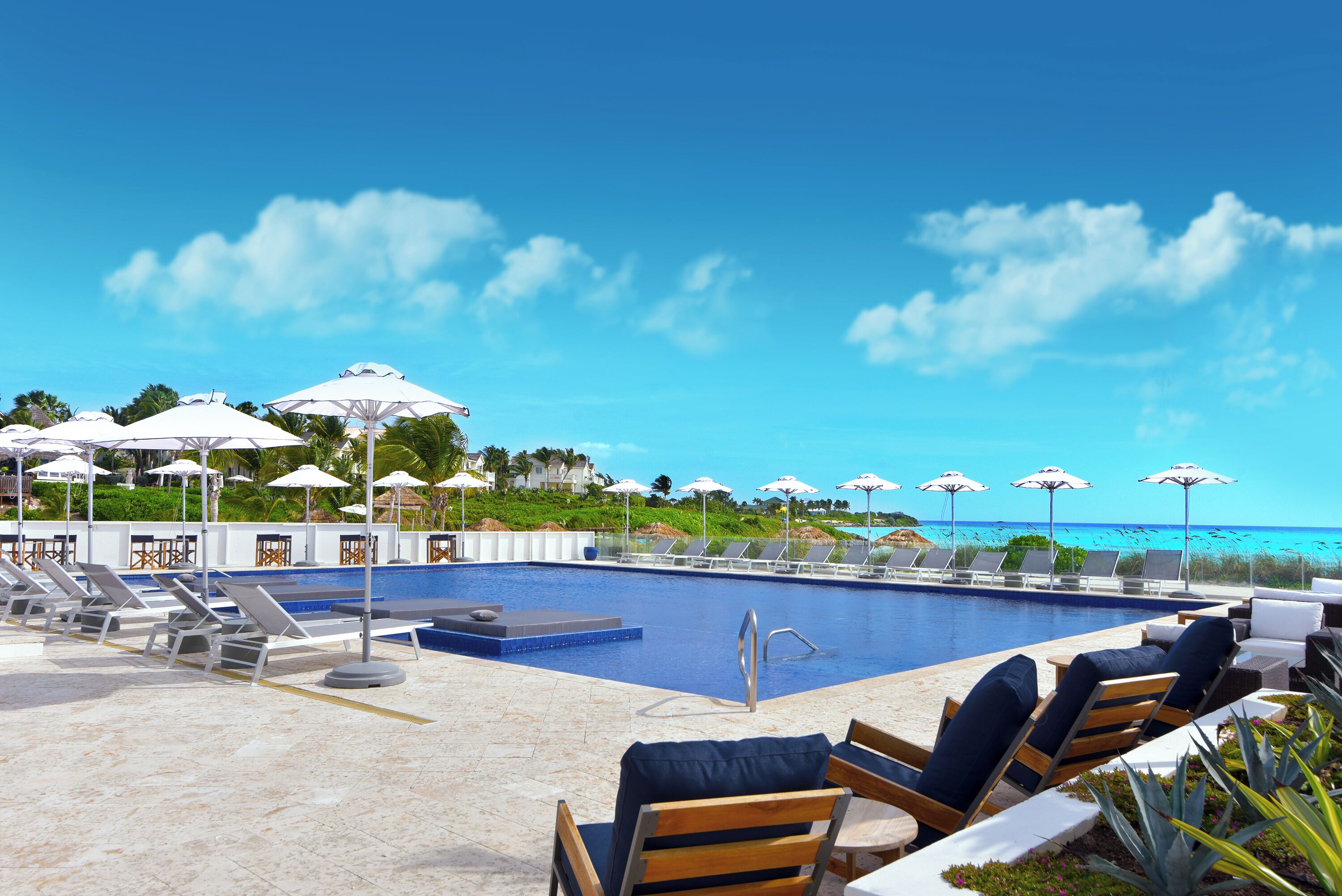 A poolside area with lounge chairs and umbrellas, a clear view of the ocean, and a pool.