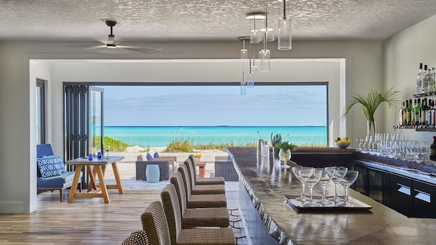 Een moderne woonkamer met een grote glazen deur die uitkijkt op het strand, een bar met glazen en flessen, en rieten meubels.