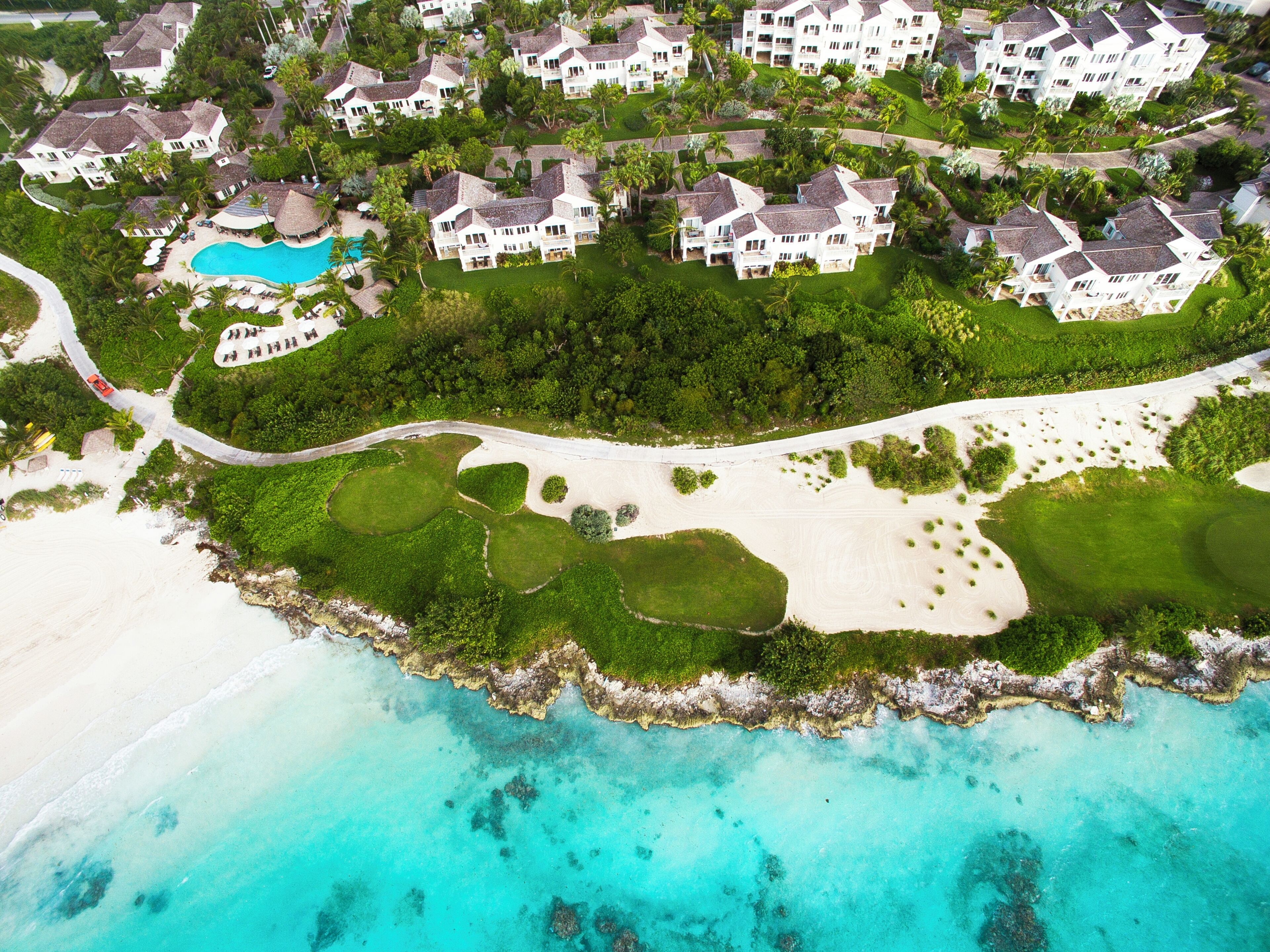 An aerial view of a resort with multiple white buildings, a central swimming pool, and a beach area with sand and turquoise waters.