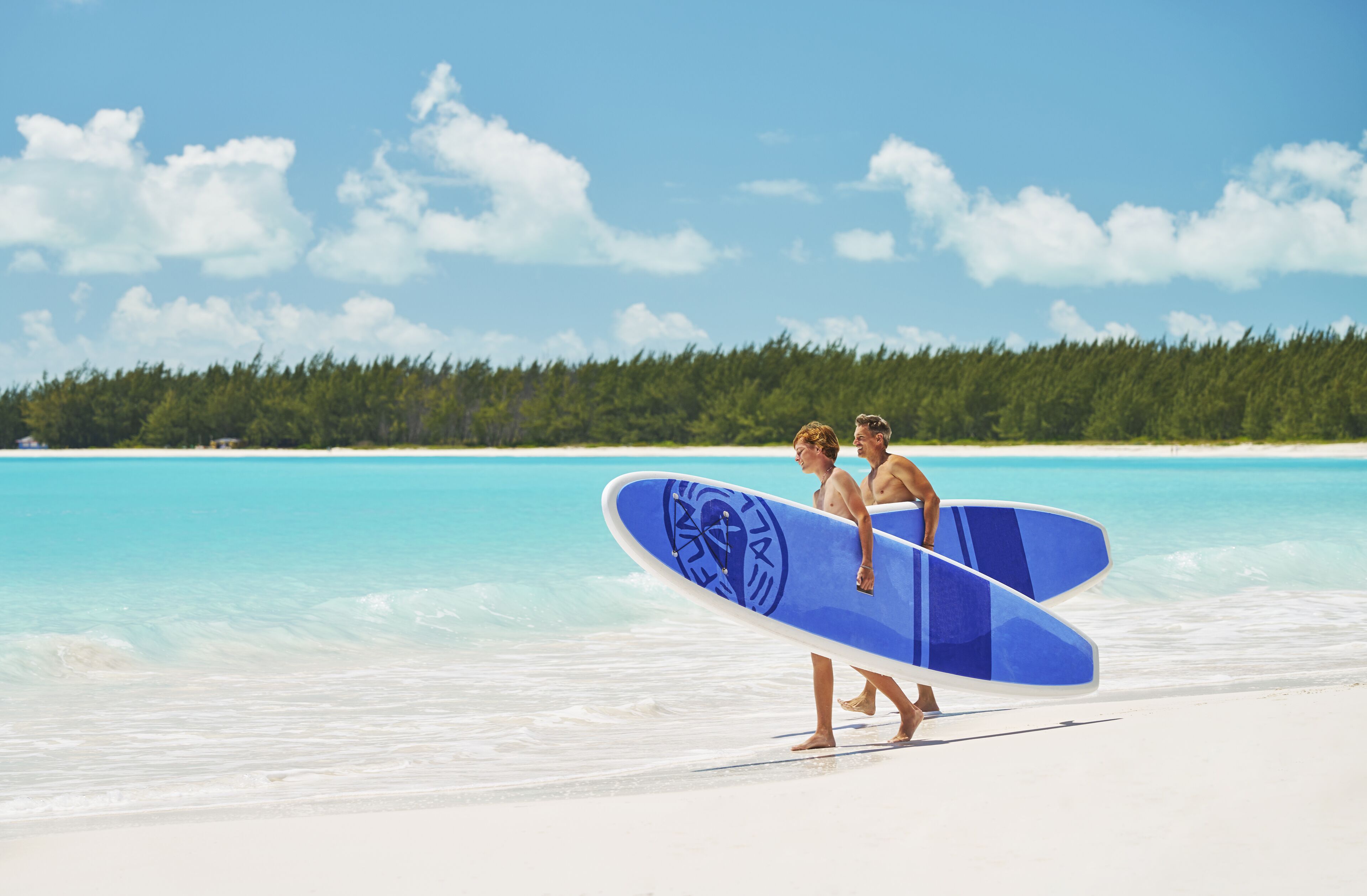 A beach scene with two people carrying a blue surfboard.