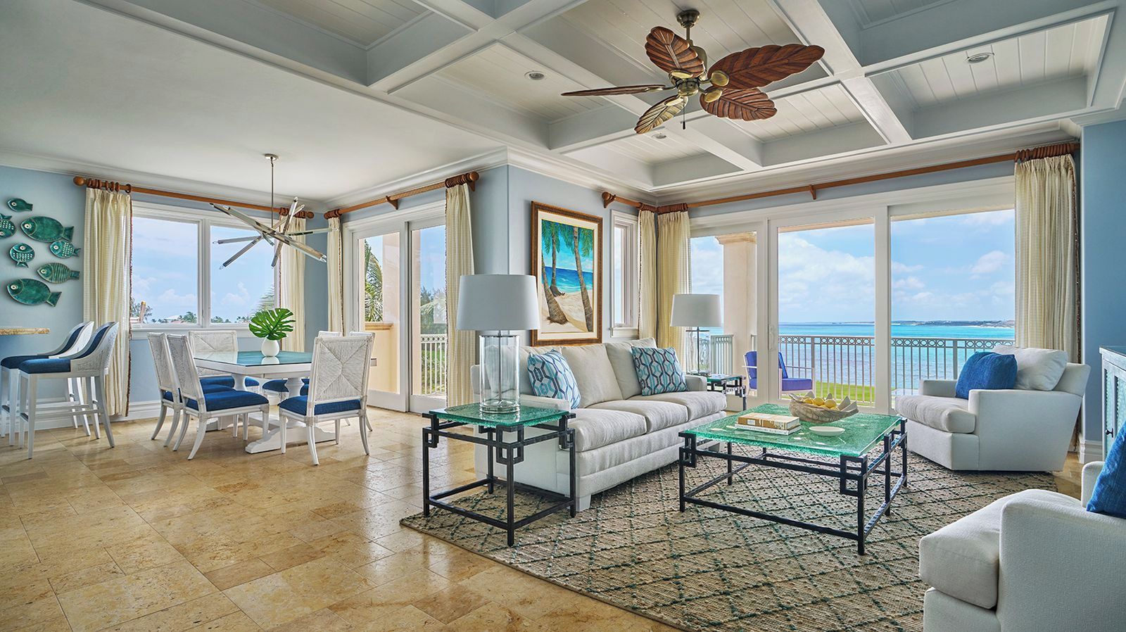 A spacious living room with a large window overlooking the ocean, a ceiling fan, and a dining area with chairs and a table.
