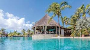 Een zwembadgedeelte van een resort met een rieten dak, palmbomen en helderblauw water.