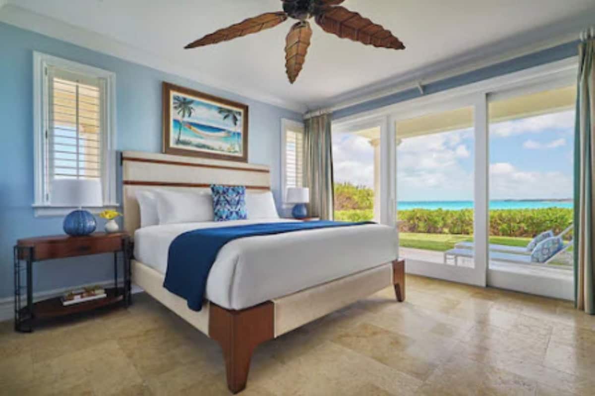 A bedroom with a bed, a nightstand, a ceiling fan, a window with a view of the ocean, and a door leading to a balcony.