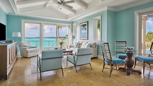 Een ruime woonkamer met een groot raam met uitzicht op een turquoise zee, een plafondventilator en uitzicht op de oceaan.