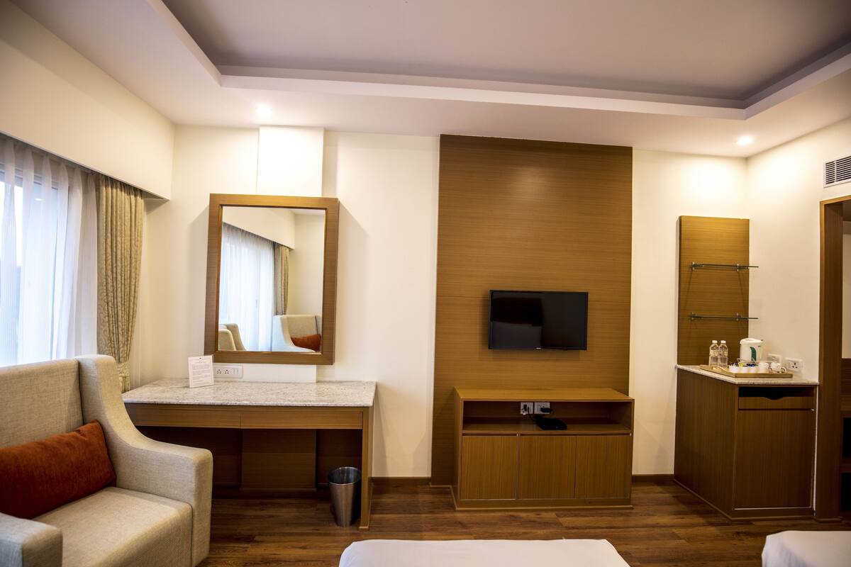 A hotel room with a bed, a sofa, a desk, a mirror, a television, a wooden cabinet, and a trash bin.