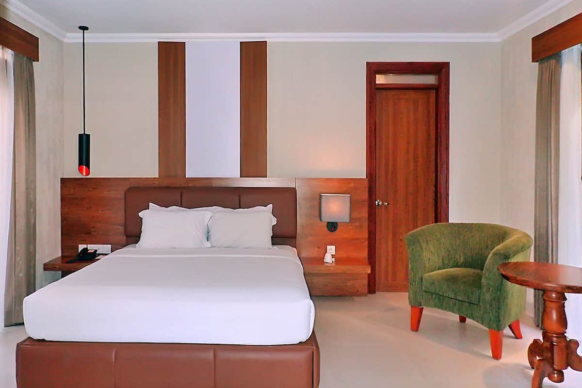 Habitación de hotel con una cama grande, un cabecero de madera, un sillón verde, una mesita de madera y una lámpara de pared.