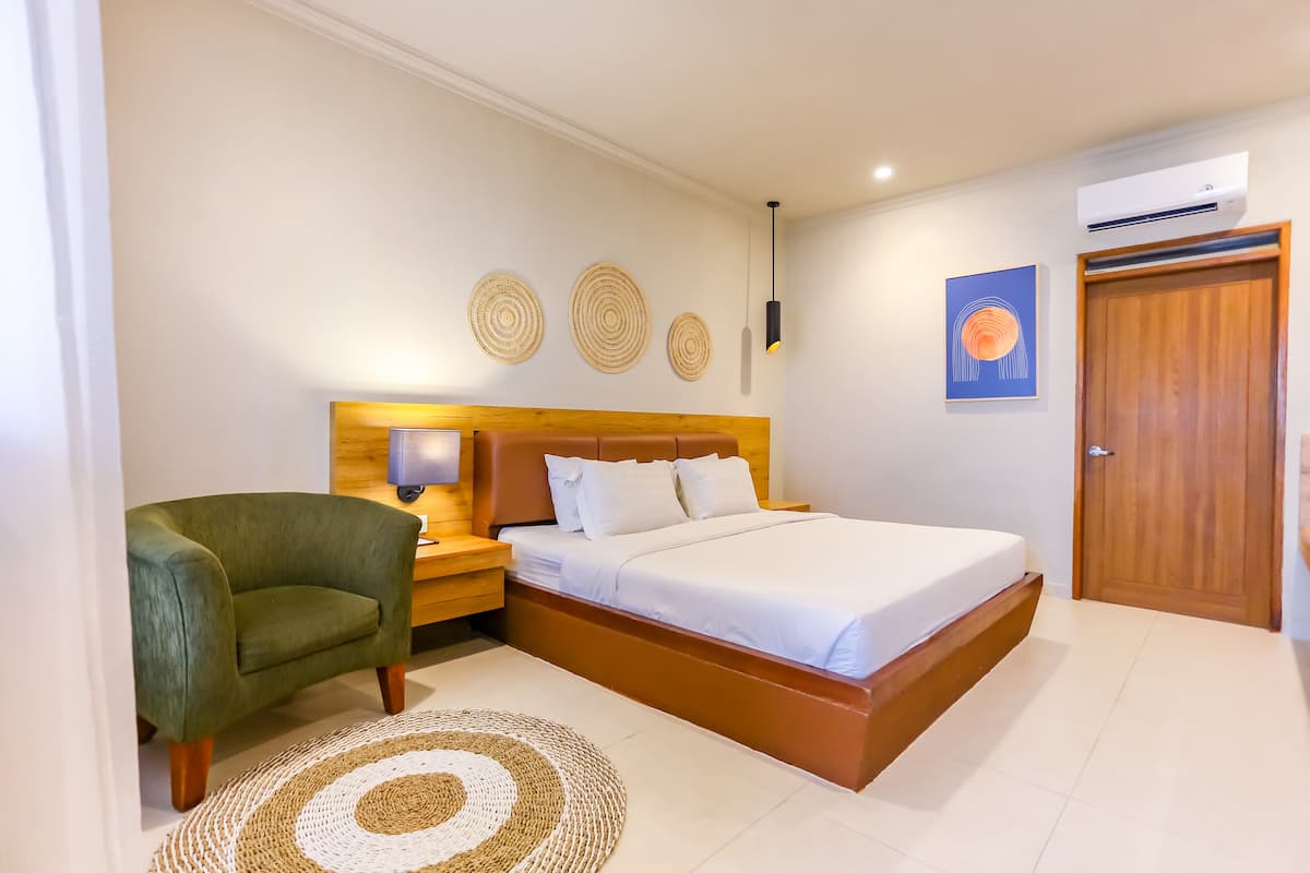 Un dormitorio ordenado con una cama, un sillón verde, una mesita de noche y un aire acondicionado de pared.