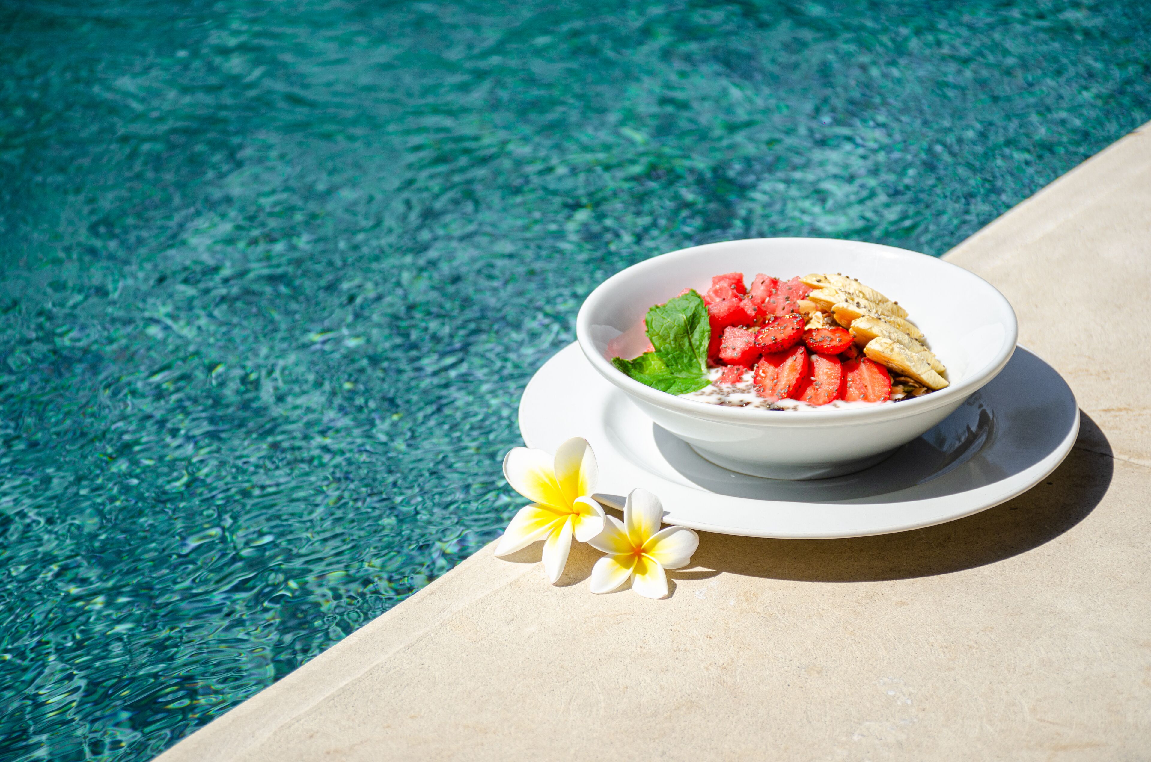 A bowl of food with a slice of watermelon and mint leaves on a white plate, placed on a poolside surface.