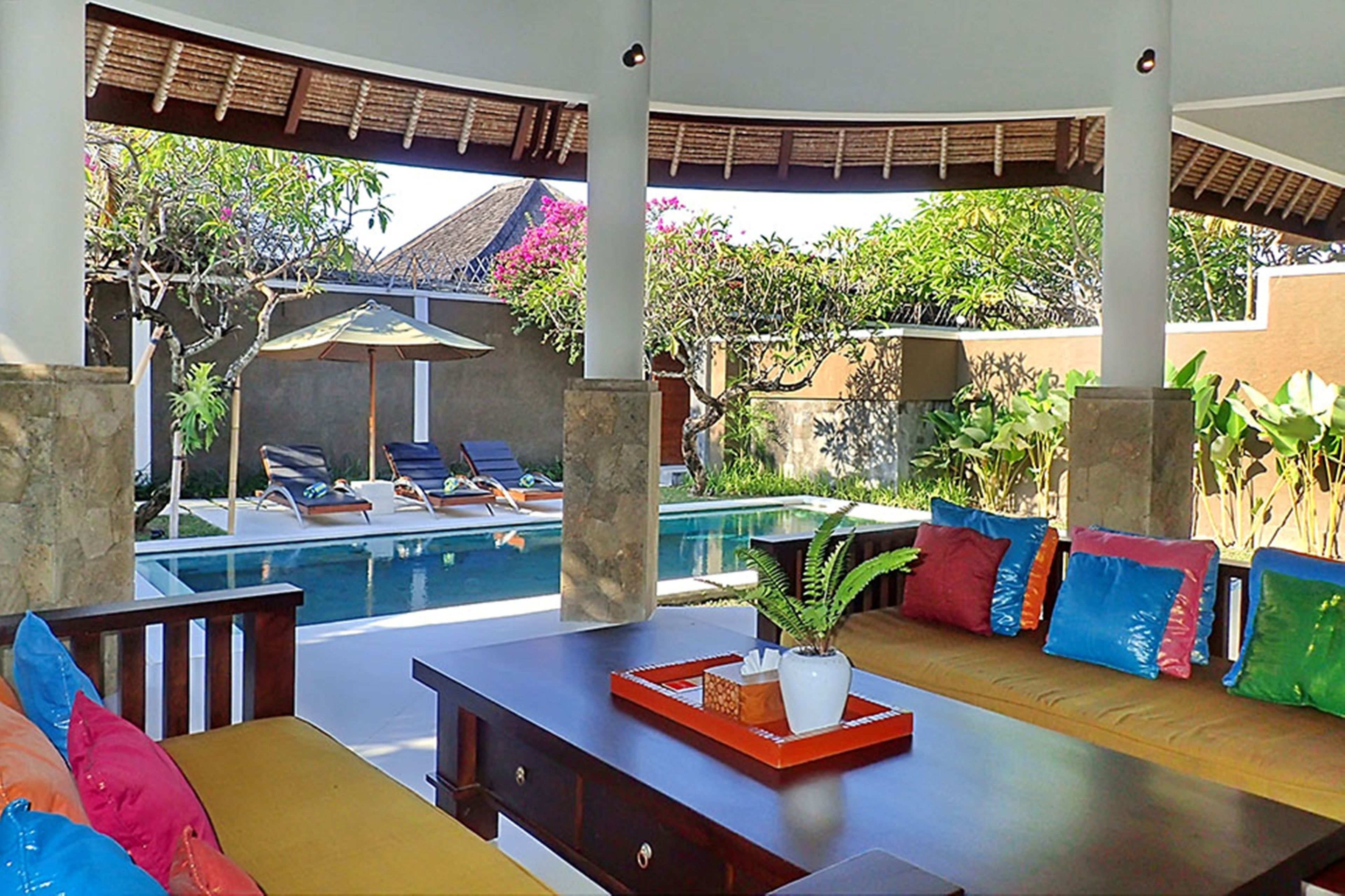 Un espace aménagé au bord de la piscine, avec un banc en bois, des coussins colorés et une table sur laquelle se trouvent un plateau et une plante.