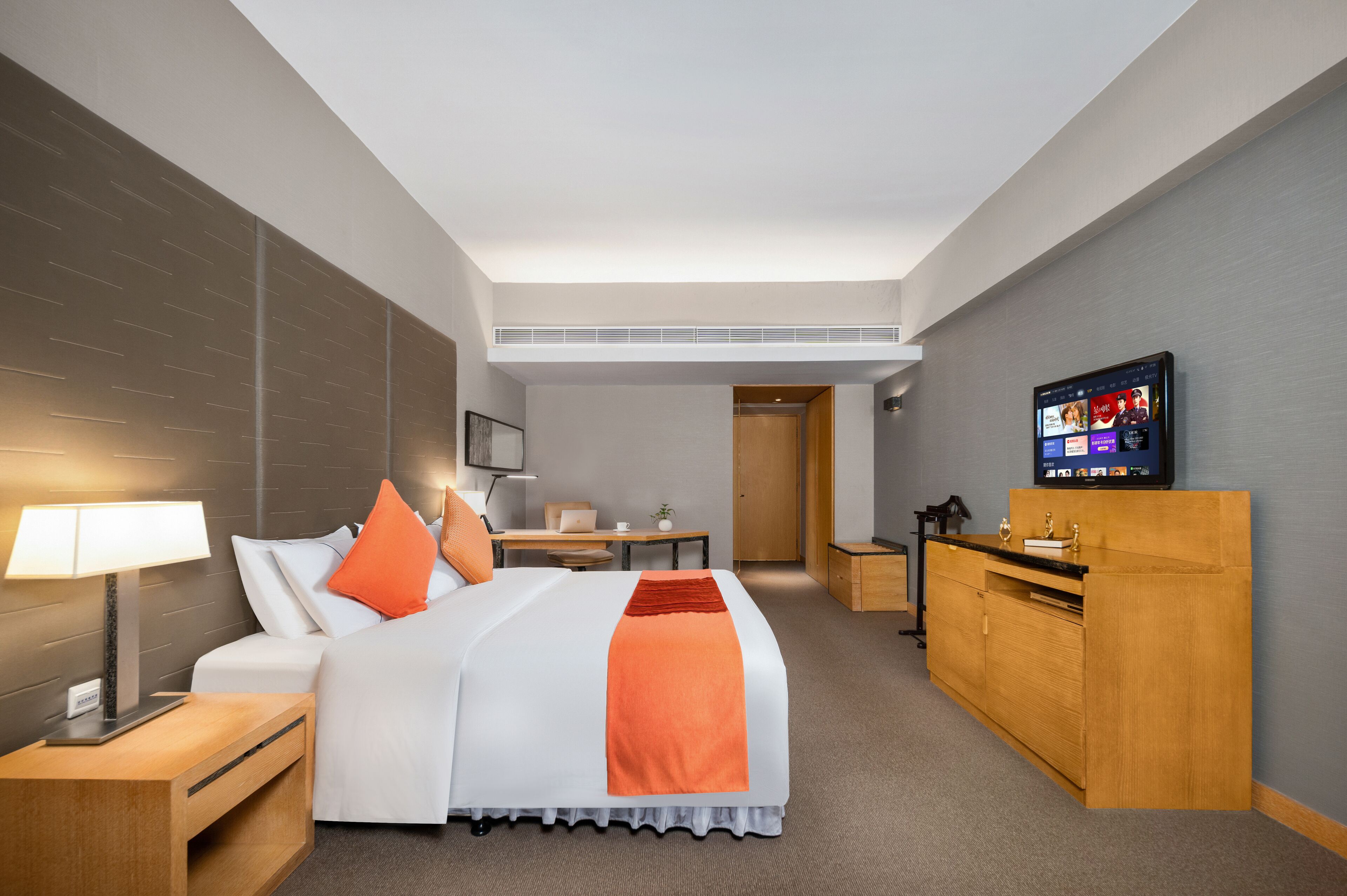 Quarto de hotel com cama, mesa de cabeceira, abajur, televisão, escrivaninha, cadeira e guarda-roupa.