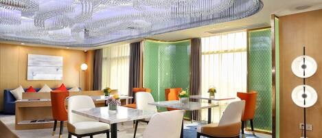 Quarto de hotel moderno com um lustre grande, área de refeições com mesa e cadeiras, e área de estar com sofás e mesa de centro.