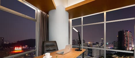 A modern hotel room with a large window, a desk with a laptop, a chair, and a lamp.