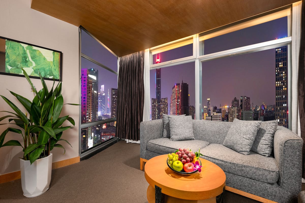 Een moderne hotelkamer met een grijze bank, een ronde houten tafel met fruit, een groot raam met uitzicht op de stad en een potplant.