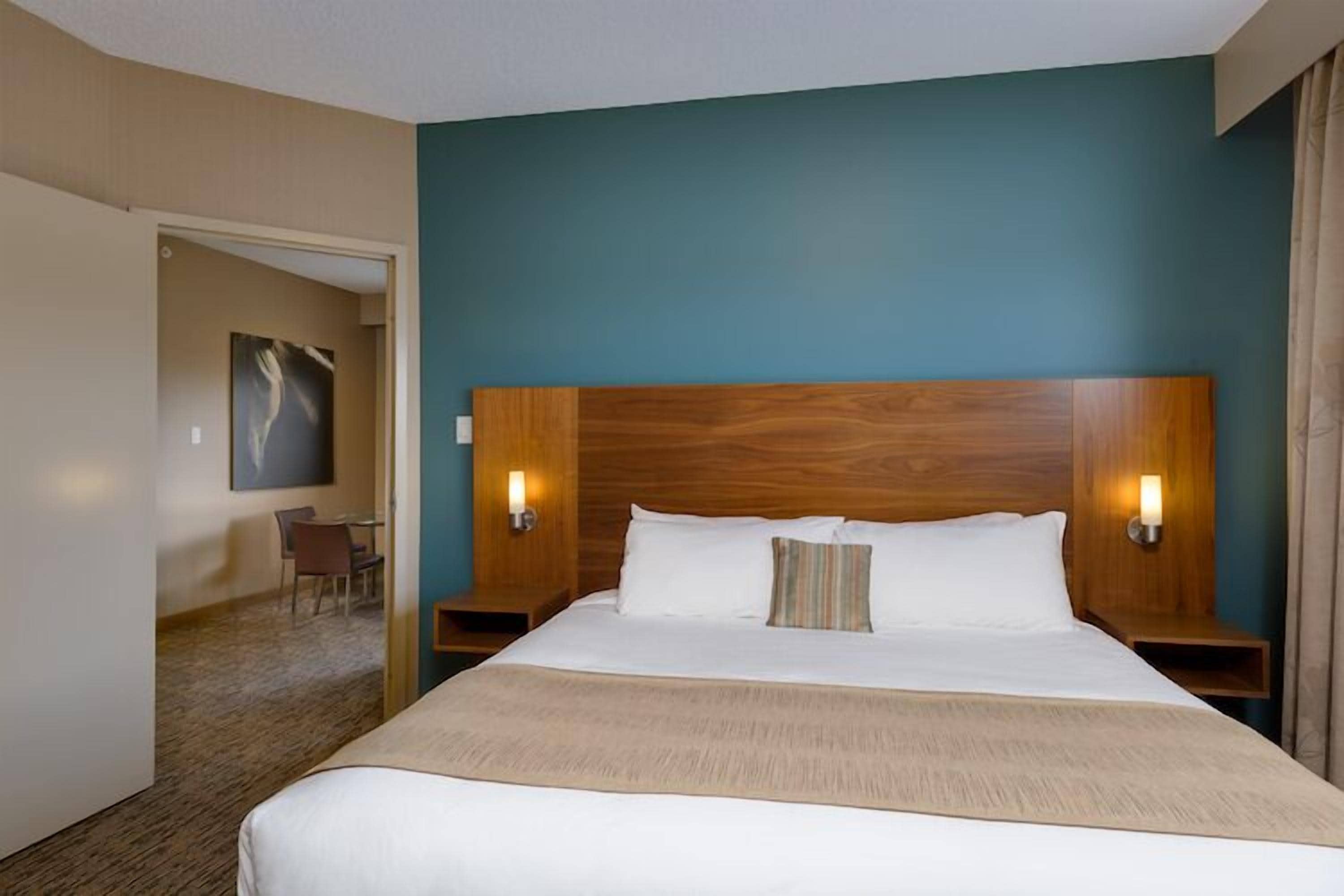 A hotel room with a large bed, wooden headboard, two bedside lamps, and a view of a dining area.