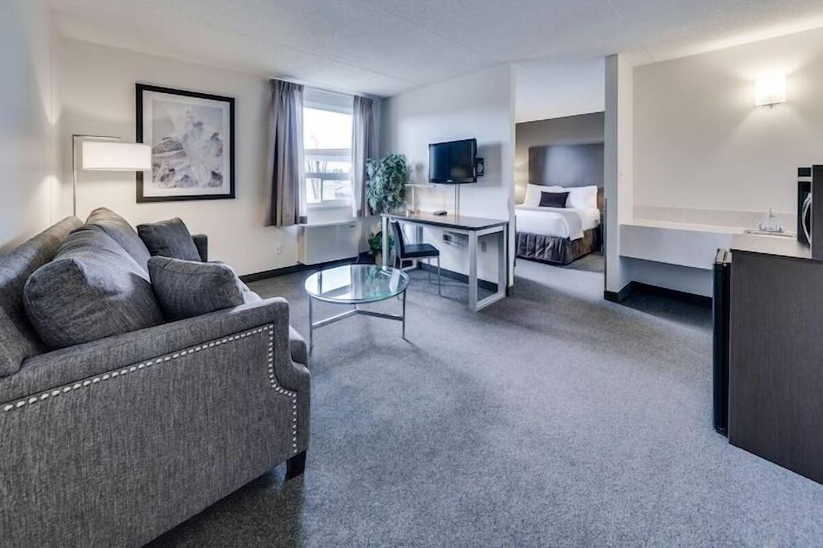 A hotel room with a grey sofa, a small round table, a bed with a white headboard, a TV, and a desk with a chair.