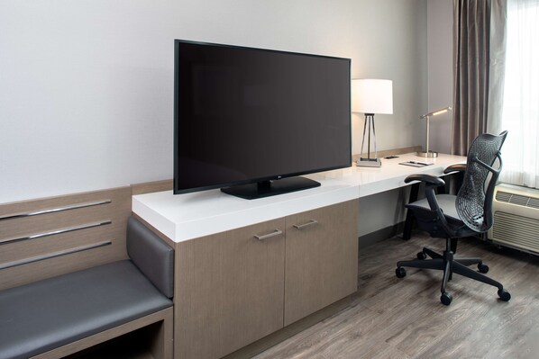 A modern hotel room with a flat-screen TV, a desk with a lamp, a chair, a bench, and a window with curtains.