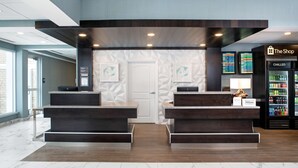 Zona de recepción de hotel con diseño moderno, que incluye un mostrador de recepción, una pantalla digital mostrando información y un refrigerador minibar.
