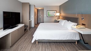 Una habitación de hotel con cama, mesita de noche, lámpara y televisor sobre un soporte.