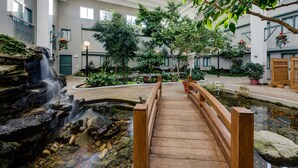 A wooden bridge over a water feature in a hotel courtyard with indoor plants and a stone wall. - Heritage Inn Hotel & Convention Centre Taber (Taber)
