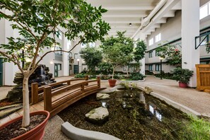 An indoor garden with a wooden bridge over a pond, surrounded by potted plants and trees. - Heritage Inn Hotel & Convention Centre Taber (Taber)