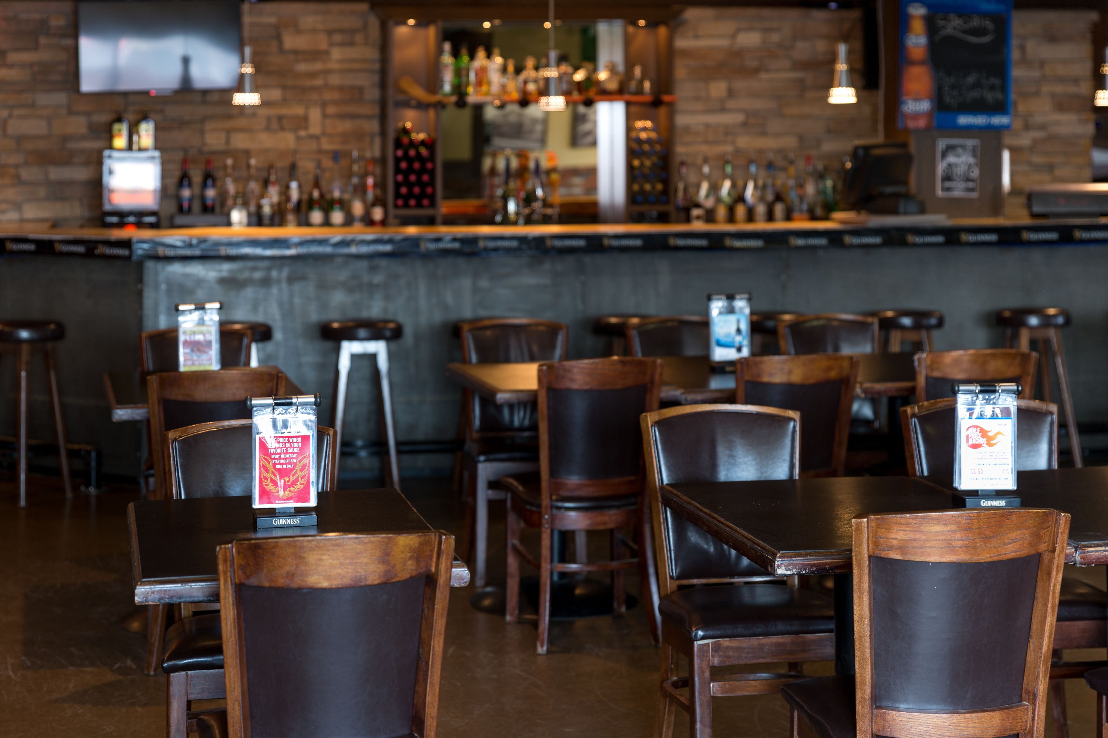 A bar area with multiple stools and tables, a brick wall, and shelves with bottles.