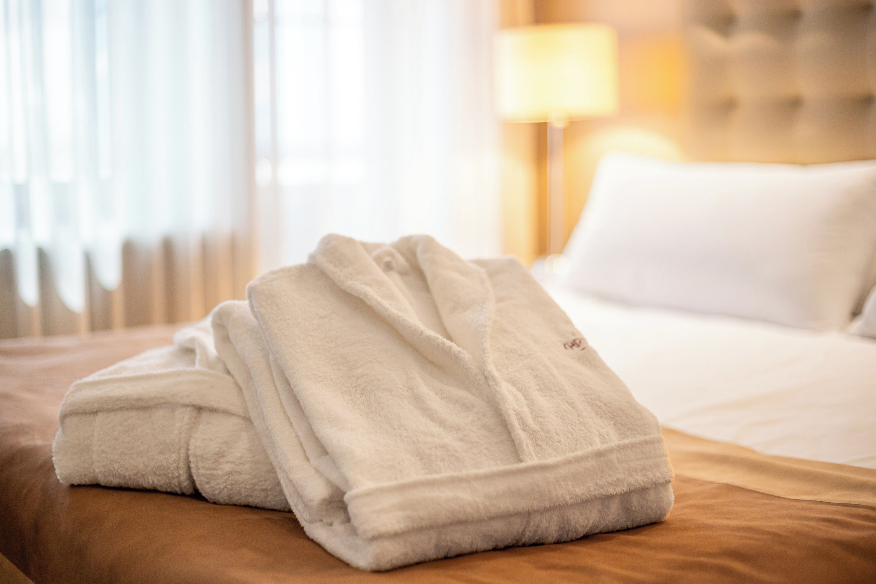 A neatly folded white bathrobe on a bed with a headboard and a lamp in the background.