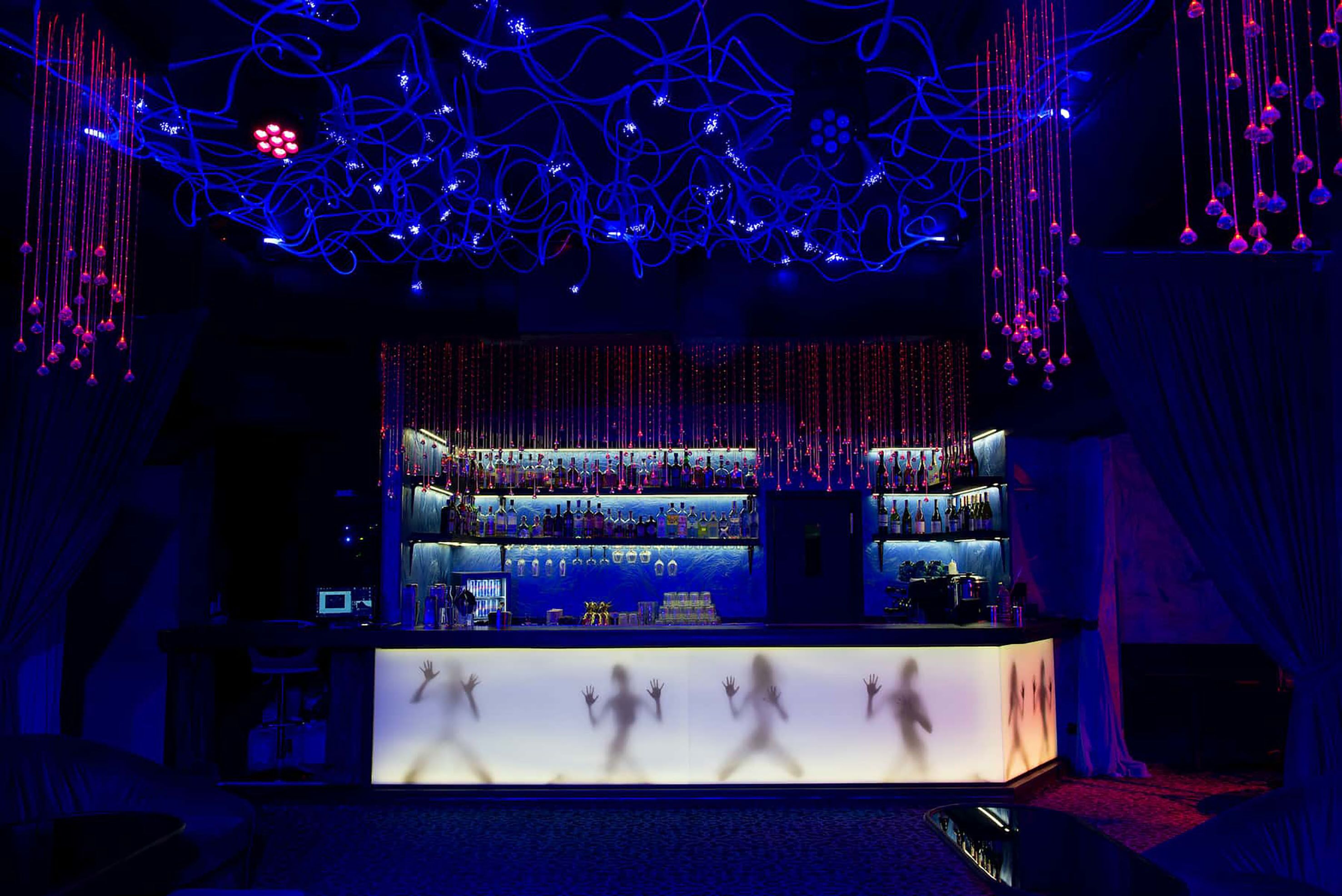 A bar area with a counter, shelves of bottles, and hanging lights.
