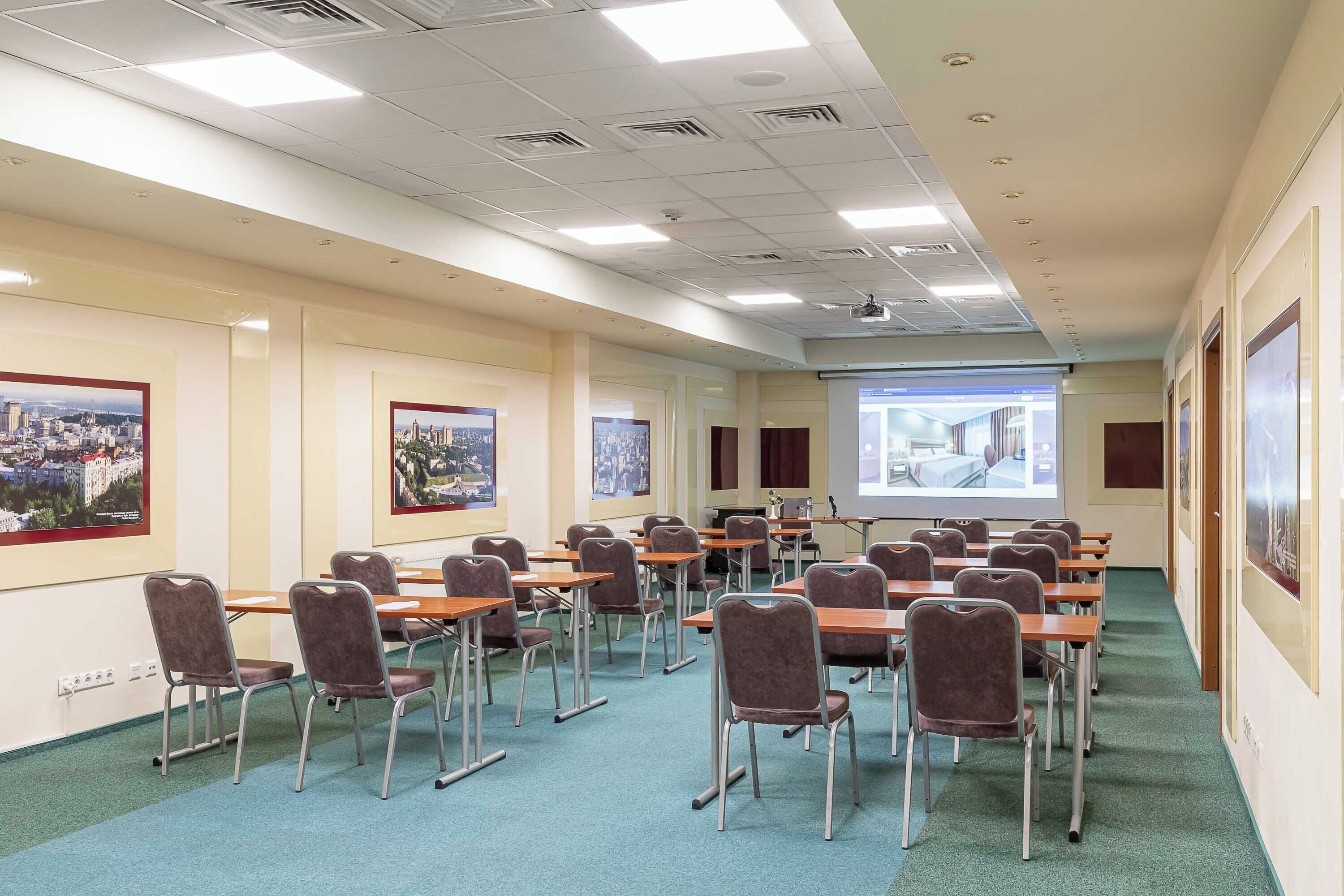 A conference room with rows of tables and chairs, a projector screen, and cityscape posters on the walls.