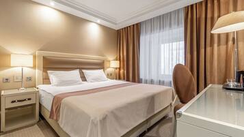Habitación de hotel con una cama grande, dos mesitas de noche con lámparas, un escritorio con silla y una ventana con cortinas.