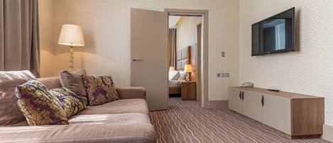Quarto de hotel com sofá, TV fixada na parede, mesa de cabeceira com abajur e carpete estampado.