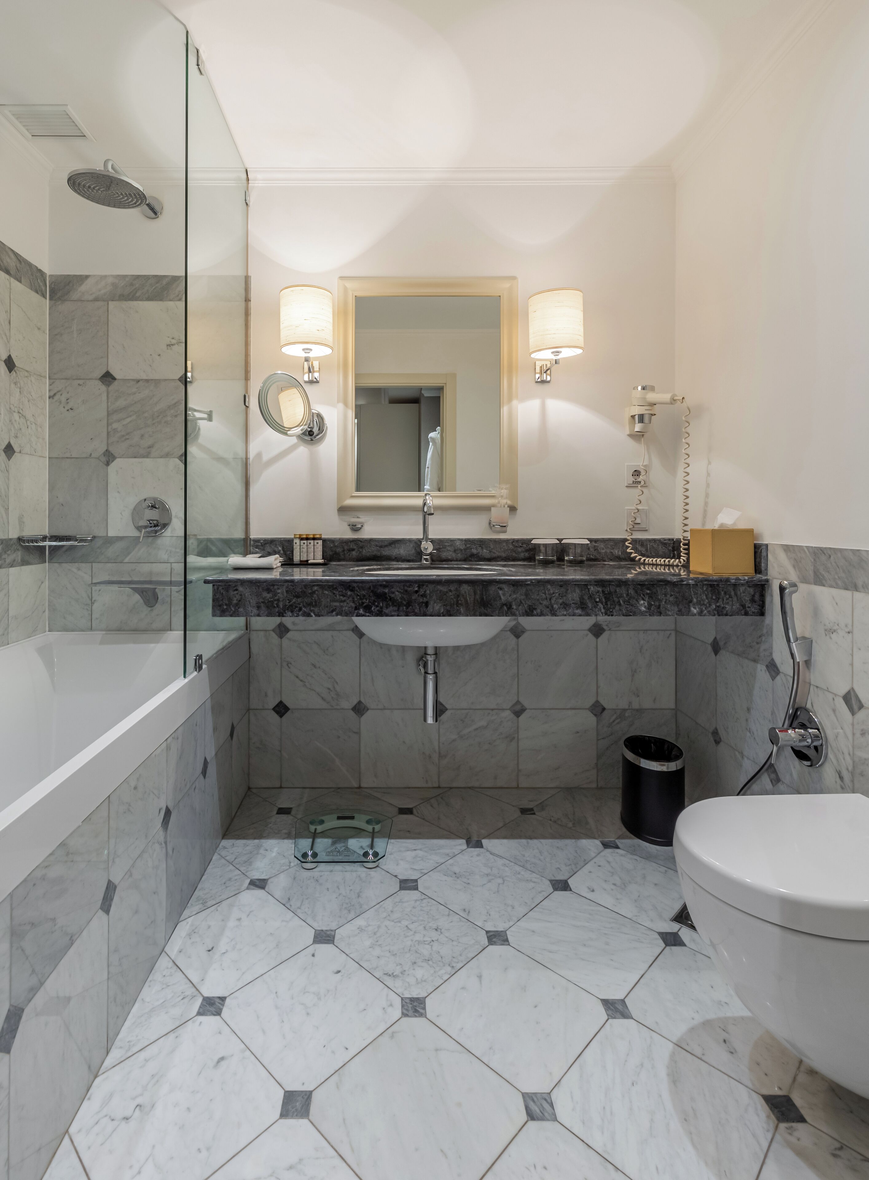 A modern bathroom with a glass-enclosed shower, a marble-tiled floor, a white sink with a mirror above, and a toilet.