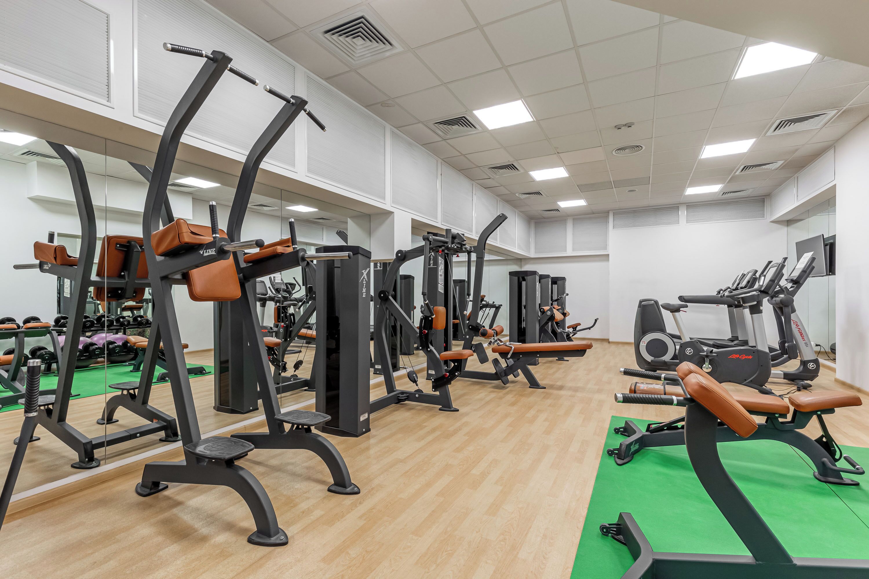 Un gimnasio con diversas máquinas de ejercicio, espejos y una zona con colchonetas verdes.