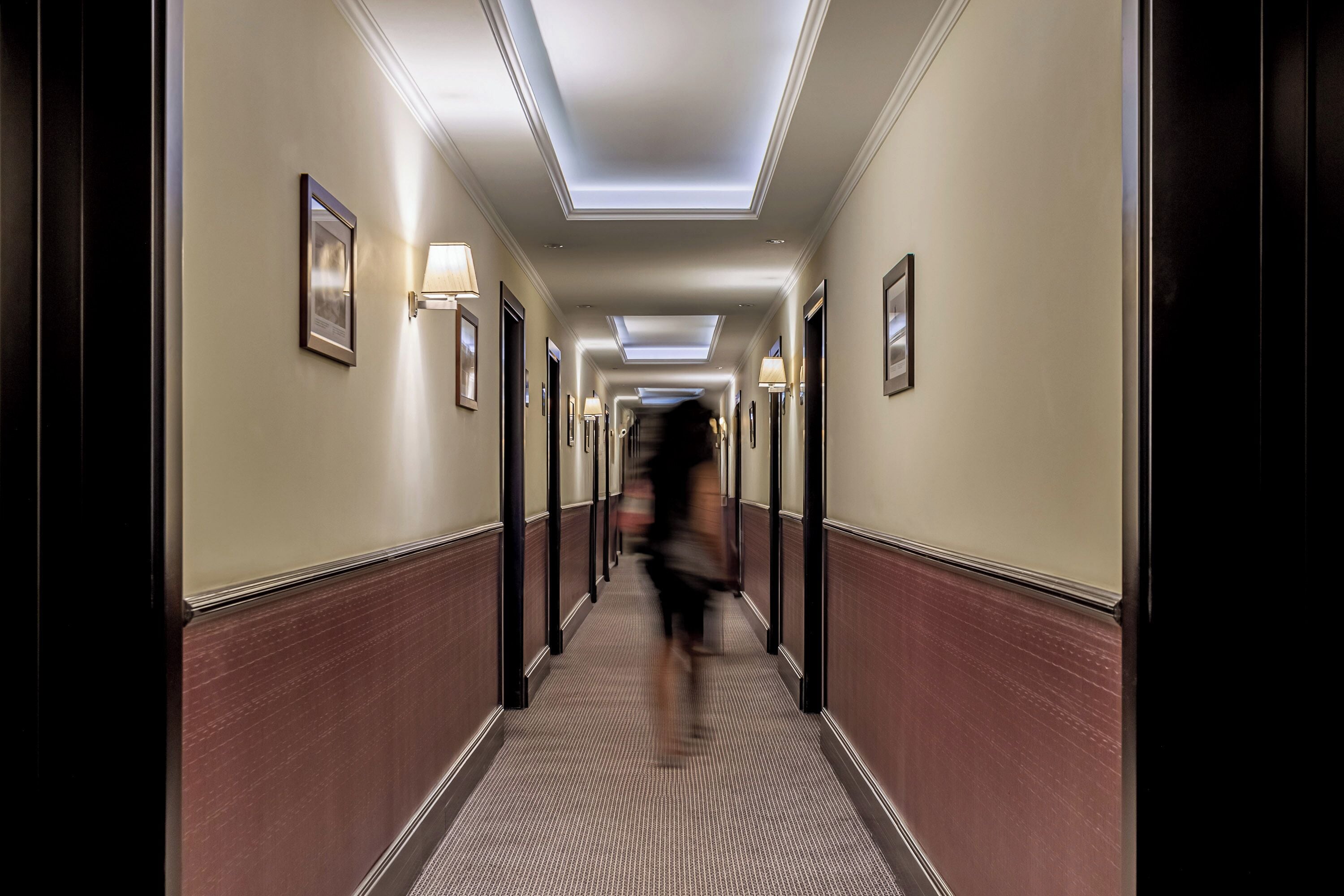 Un pasillo de hotel con un candelabro central, cuadros enmarcados en las paredes y una alfombra estampada.