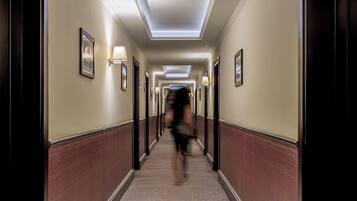 A hotel hallway with a central light fixture, framed pictures on the walls, and a patterned carpet.