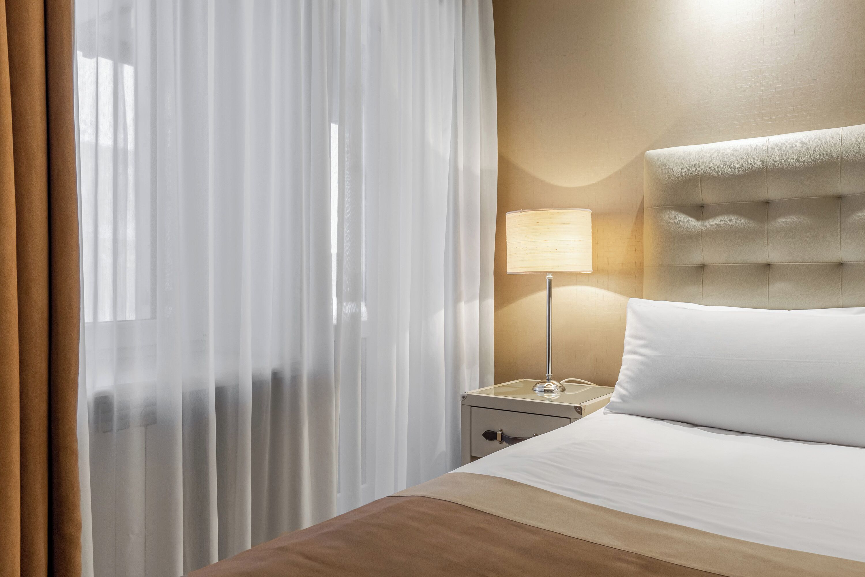 A hotel room with a bed, a nightstand, a lamp, and sheer curtains.