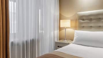 Quarto de hotel com cama, mesa de cabeceira, abajur e cortinas translúcidas.