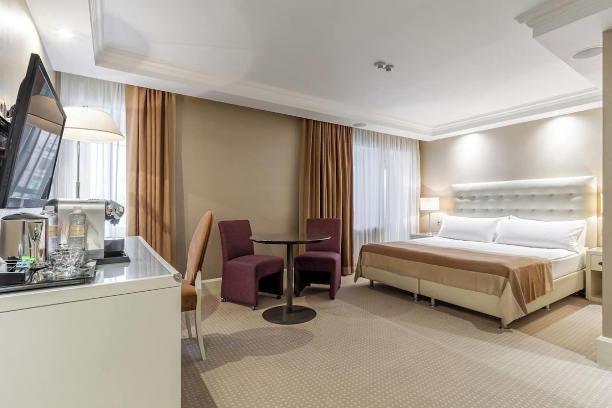 A hotel room with a bed, a desk with a lamp, two chairs, and a television.