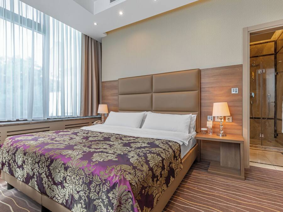 Habitación de hotel con una cama grande, una mesita de noche con lámparas, zona de ducha y vistas al exterior a través de la ventana.