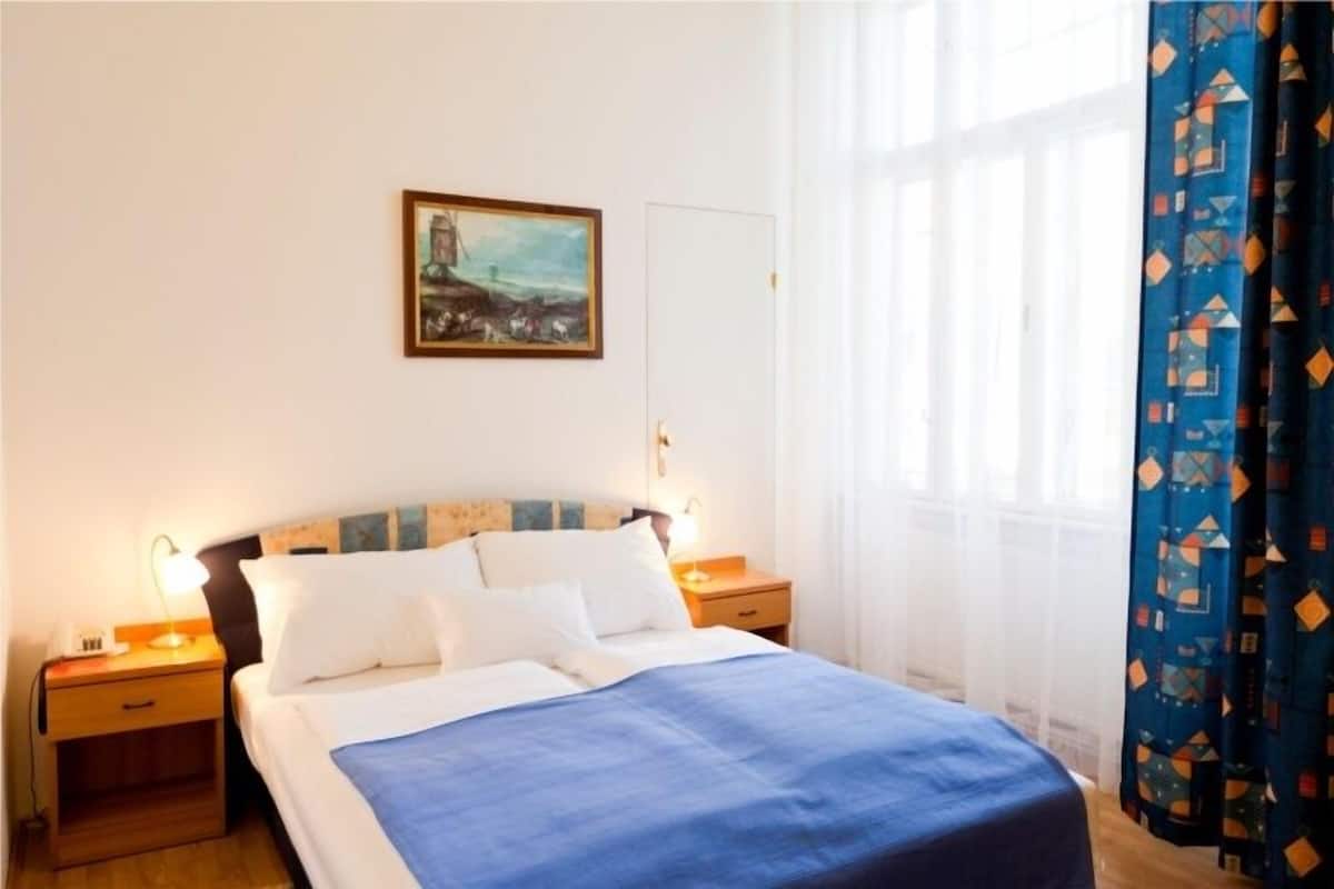 A hotel room with a bed, two bedside tables, a nightstand, a painting on the wall, and a window with curtains.