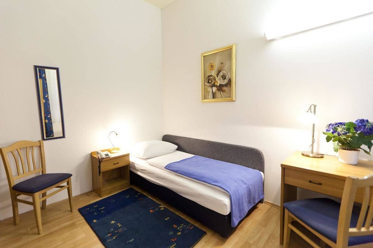 A bedroom with a bed, two chairs, a desk, a nightstand, a lamp, a potted plant, and a framed picture on the wall.