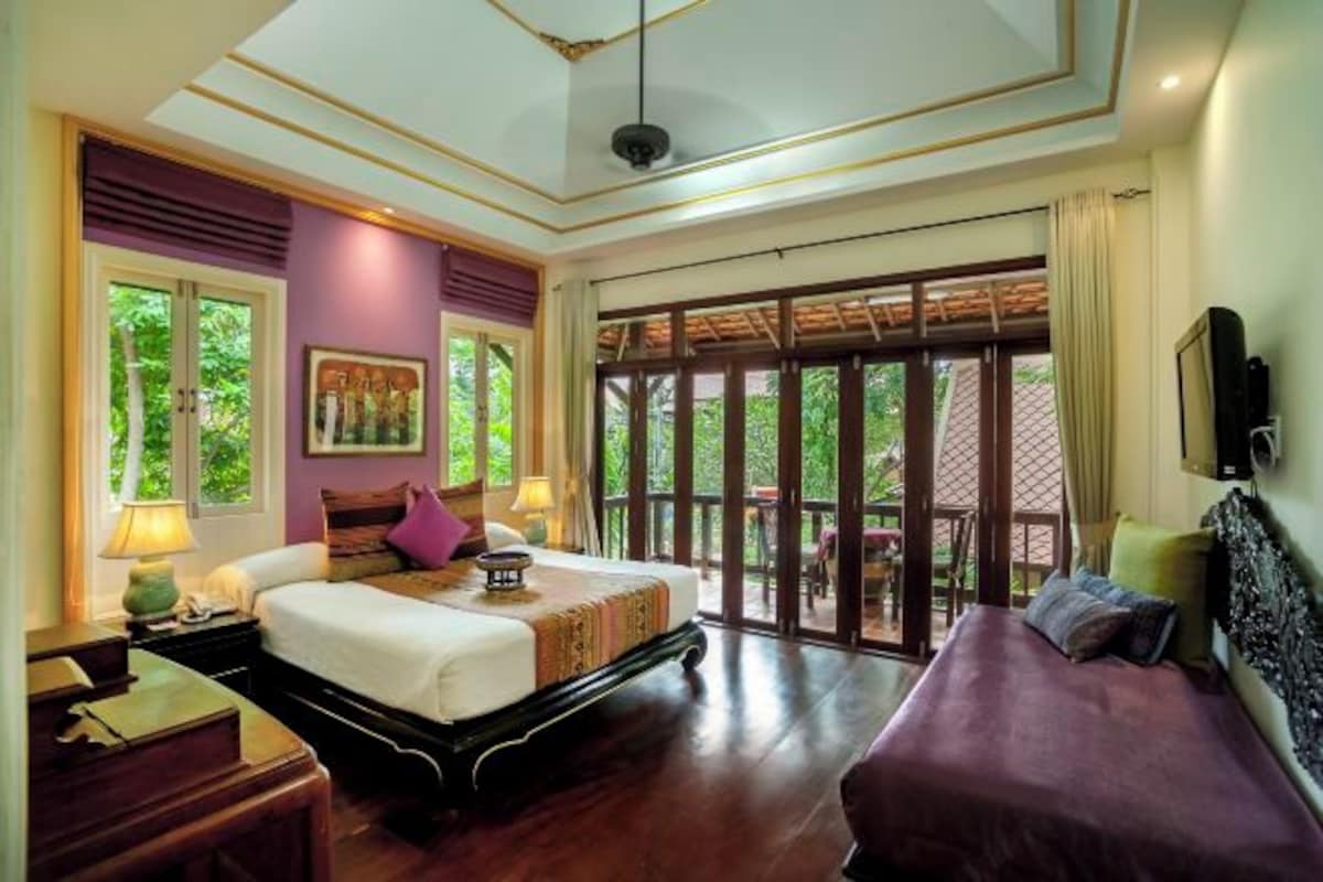 A bedroom with a large bed, a sofa, a desk, and a window with a view of greenery.