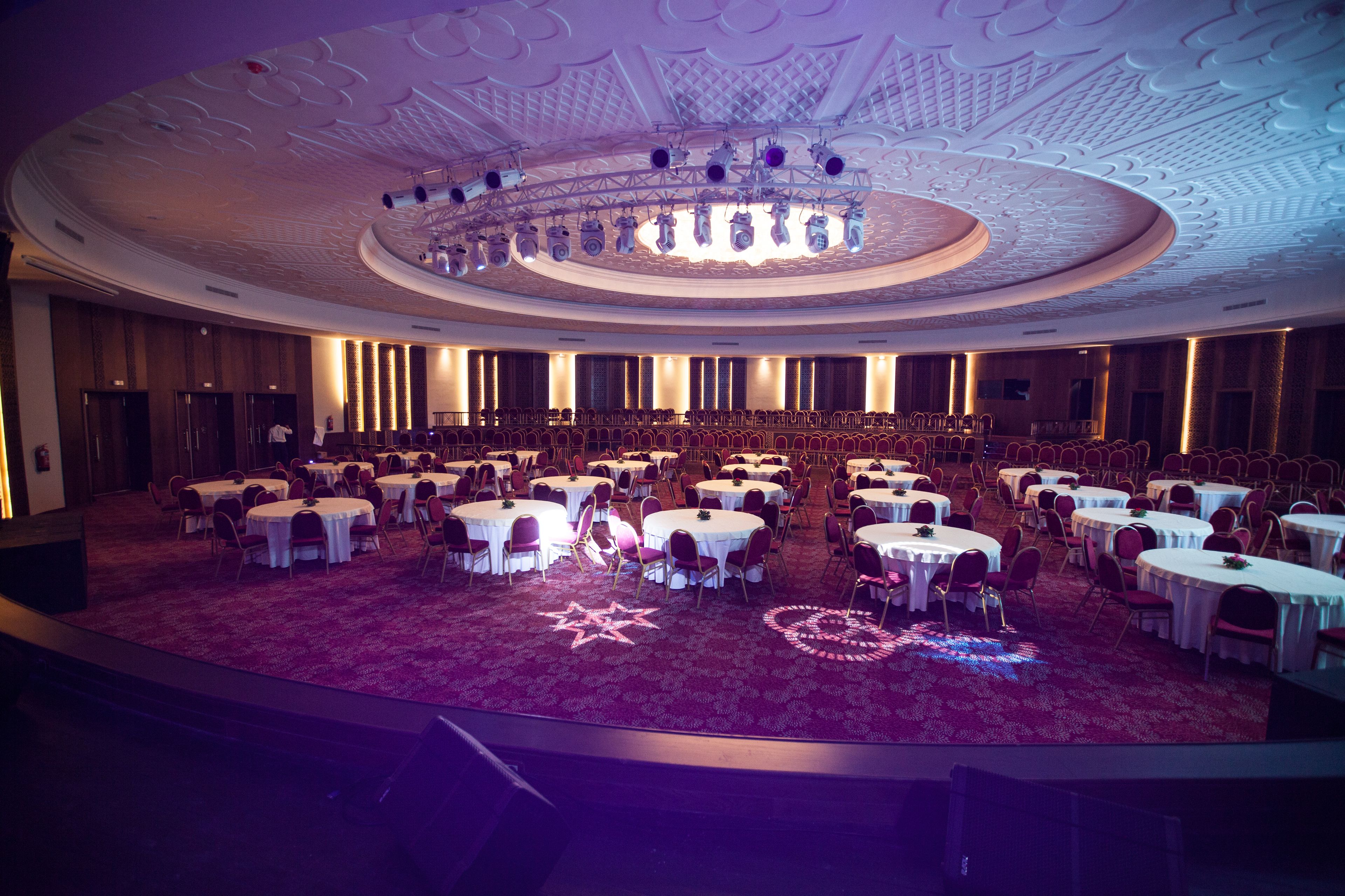 Un gran salón de baile con mesas redondas y sillas, un escenario central y un techo decorativo.