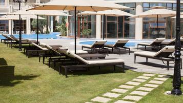 Zona junto a la piscina con sillones de descanso y sombrillas, una piscina y un edificio con grandes ventanales al fondo.