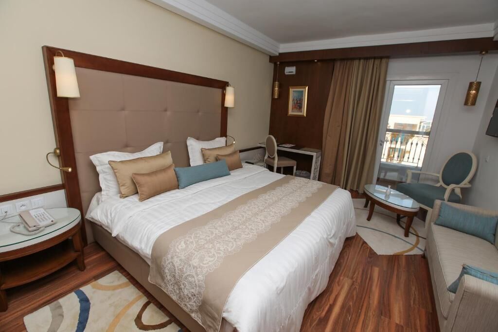 A hotel room with a large bed, bedside tables, a desk, a chair, a sofa, and a balcony with a view.