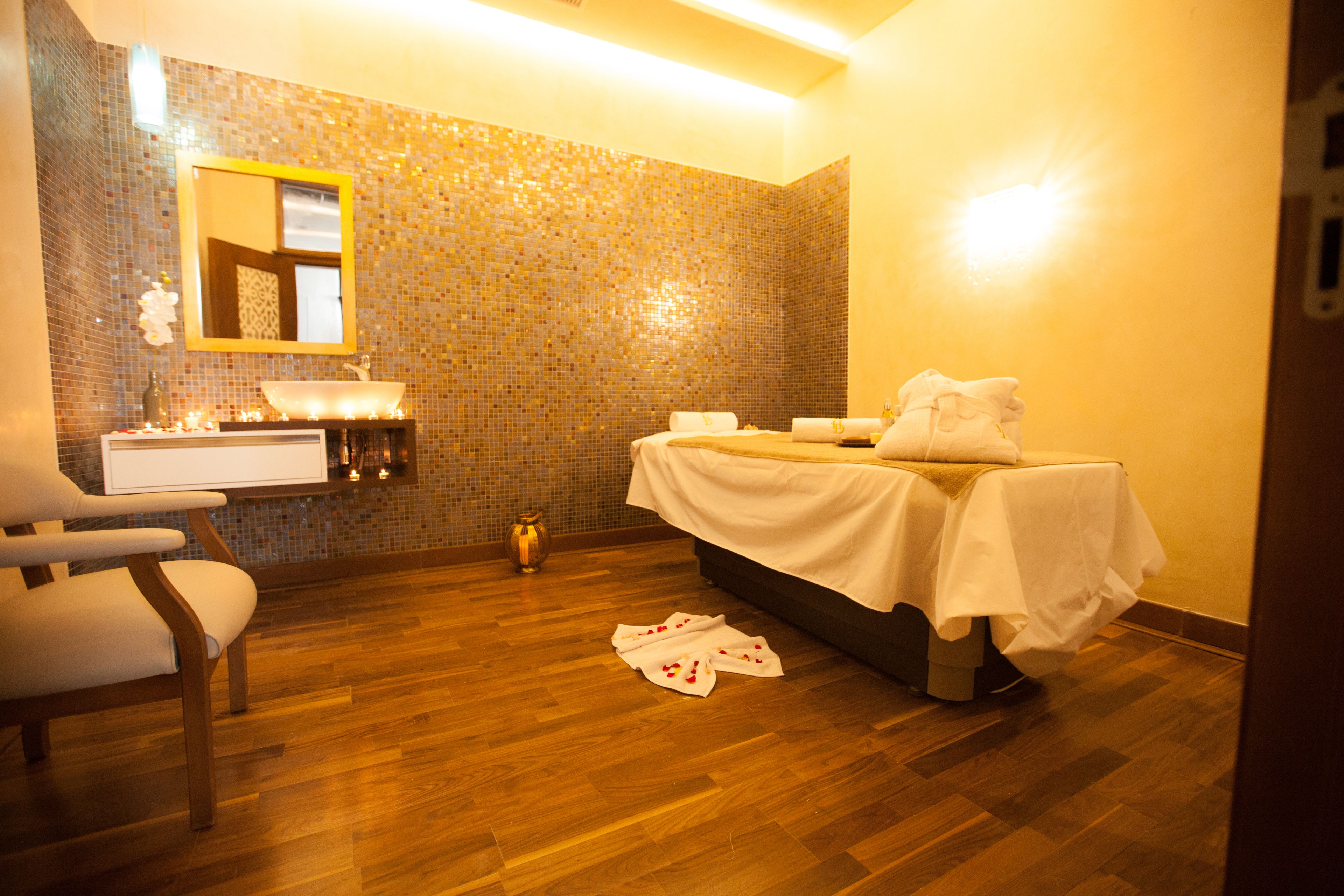 A room with a wooden floor, a white bed with a white cover, a white sink, a mirror, and a chair.