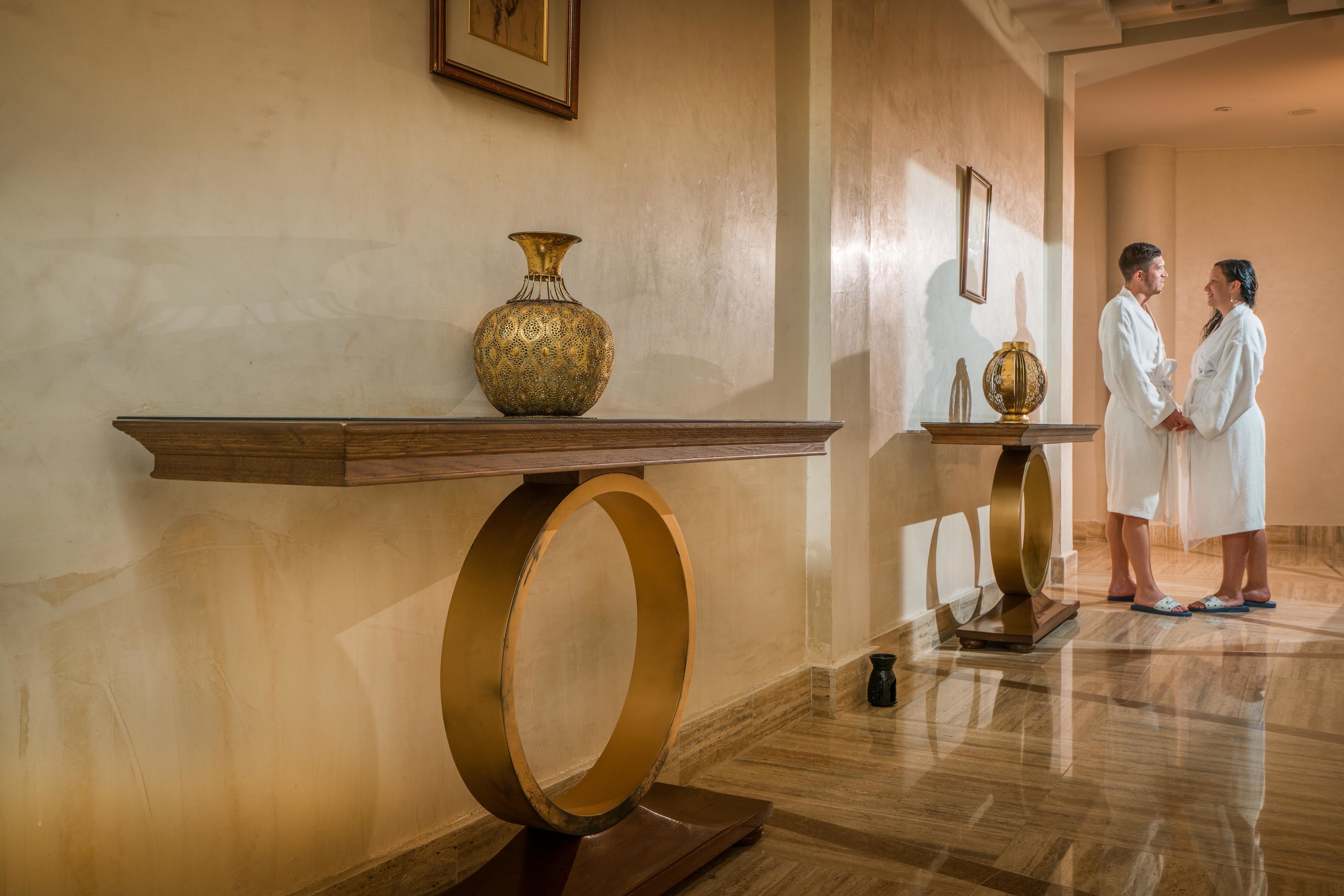 A lobby with a marble floor, a wooden console table with a brass vase, and a couple in white robes.